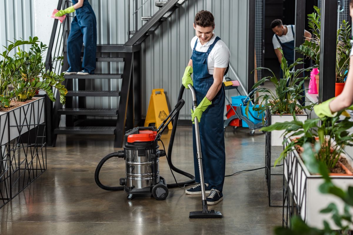 young cleaner cleaning floor with vacuum cleaner near multicultural colleagues