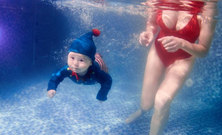 Horizontal snapshot of very interesting swimming class. Cute infant wearing elf suit, mother wearing red swimming suit, swimming underwater in pool. Baby holding breath. Concept of active lifestyle