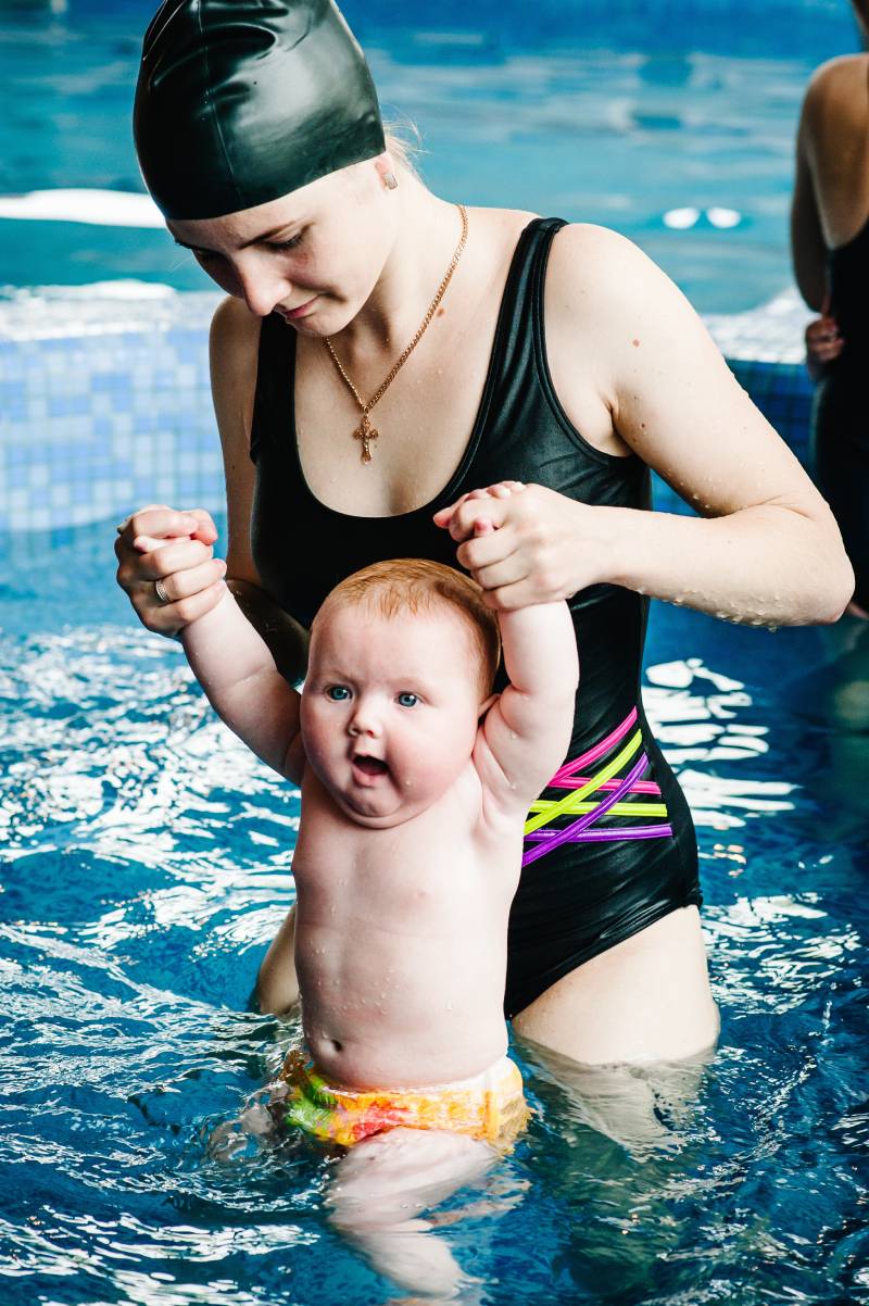 Young mother, swimming instructor and happy little girl in paddling pool. Teaches infant child to swim. Enjoy first day of swimming in water. Mom holds hand child preparing for diving. doing exercises