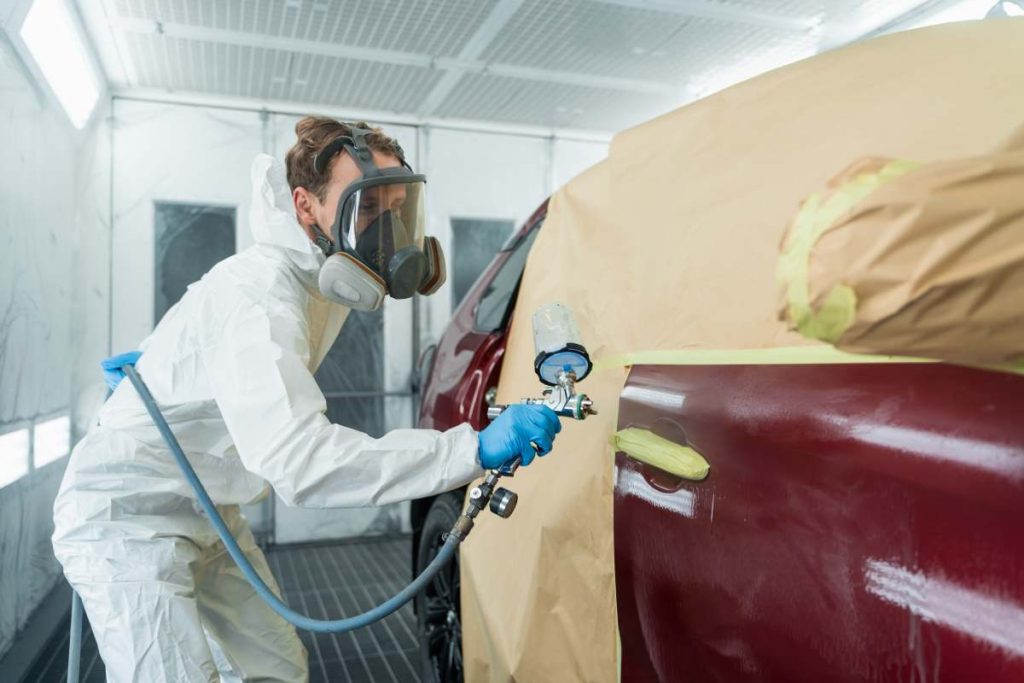 Auto painter in a protective suit and mask sprays a degreaser or other cleaner on a car door using a spray gun. Preparation of vehicle body elements for local repair