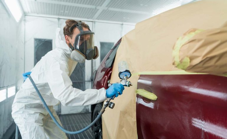 Auto painter in a protective suit and mask sprays a degreaser or other cleaner on a car door using a spray gun. Preparation of vehicle body elements for local repair