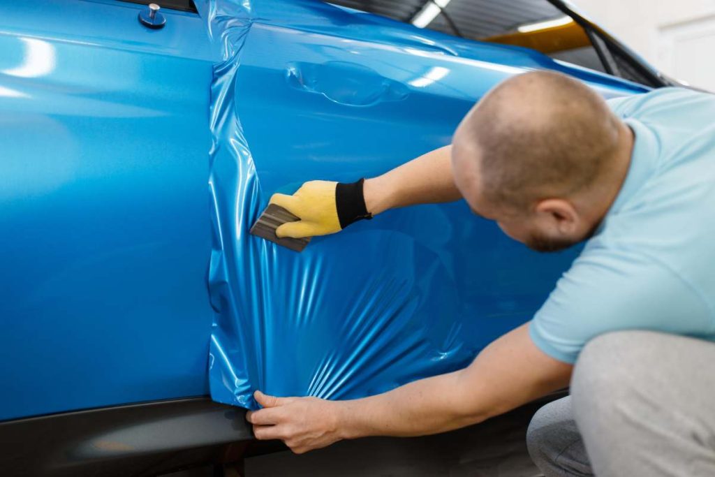 Male mechanic installs protective vinyl foil or film on vehicle door. Worker makes auto detailing. Automobile paint protection, professional tuning